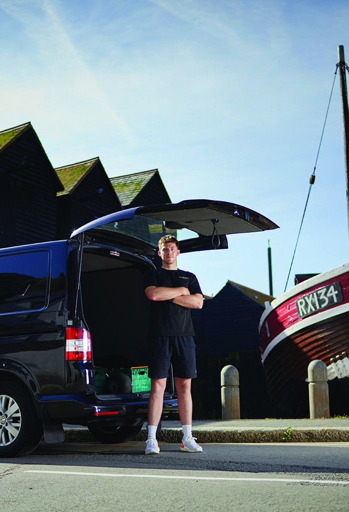Adam Shaw standing at the open tailgate of his black van on the Stade at Hastings Old Town, with the black net huts and the fishing boat RX134 behind him